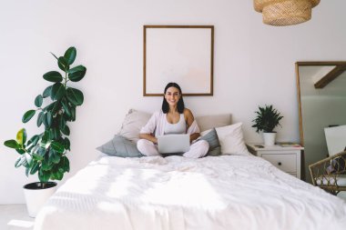 Keyifli genç bayan serbest çalışan rahat bir yatakta oturuyor ve evdeki modern yatak odasından uzak bir iş için dizüstü bilgisayara bakıyor.
