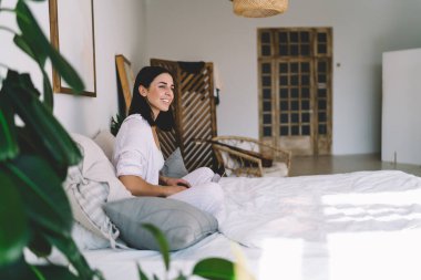 Gülümseyen genç bayan serbest çalışanın rahat yatağında dinlenmesi ve sabah boş vakitlerinde dizüstü bilgisayarını karıştırırken başka tarafa bakması.