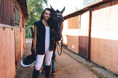 Heyecanlı genç bayan binici, uzun siyah saçlı, yelek ve botlarıyla gülümseyen ve kameraya bakan çiftlikteki ahırlarda itaatkar, koyu renk at sürücüsü.