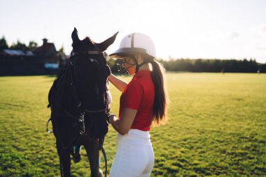Kask takan genç bayan jokey ve kırmızı polo tişörtü antrenman sırasında bulutsuz gökyüzüne karşı kestane atının yanında duruyordu.