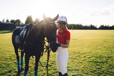 Kasklı ve üniformalı bir kadın binici. Yazın yeşil alanda bulutsuz gökyüzüne karşı antrenmandan önce kestane atının yanında duruyor.