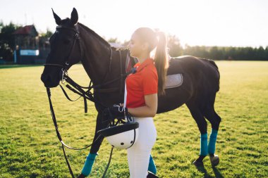 Jokey üniformalı genç kadın binicinin, güneşli bir günde çimenlik alanda bulutsuz gökyüzüne karşı kestane atının yanında duruşu.