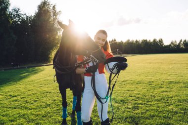 Mutlu genç atlı kadın pantolonlu ve kırmızı polo tişörtlü, gülümseyen çiftlikteki çimenli tarlada koyu renk at beslerken.