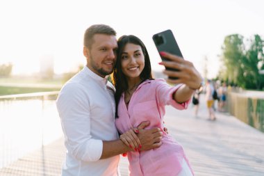 Neşeli genç çift gülümsüyor ve parkta yan yana dururken ve yazın kendi portresini çekerken akıllı telefon ekranına bakıyor.