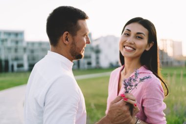 Beyaz tişörtlü erkek arkadaşın yeşil şehir parkındaki randevusu sırasında kaldırım kenarında dikilirken neşeli kız arkadaşına polen çiçekleri hediye edişi.