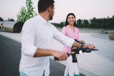 Hafta sonu gezisi boyunca elektrikli scooterlarla gezerken günlük giysiler içinde gülümseyen ve erkek arkadaşına bakan etnik bir kadın.