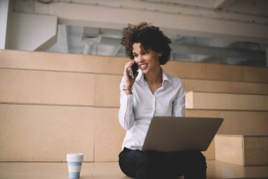 Bilgisayarlı, cep telefonuyla konuşan bin yıllık beyaz bir kız. Modern başarılı kadın kavramı. Genç, gülümseyen kıvırcık kadın, çalışma odasında bir kahve fincanıyla ahşap bankta oturuyor.