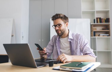 Milenyum Kafkas işadamı dizüstü bilgisayardan izliyor ve akıllı telefon kullanıyor. Modern başarılı adam kavramı. Gözlüklü genç adam açık uzay ofisindeki ahşap masada oturuyor.