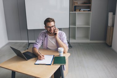Milenyum Kafkas işadamı akıllı telefonla konuşuyor ve panoya bir şeyler yazıyor. Modern başarılı adam kavramı. Gözlüklü genç adam açık uzay ofisinde masada oturuyor.