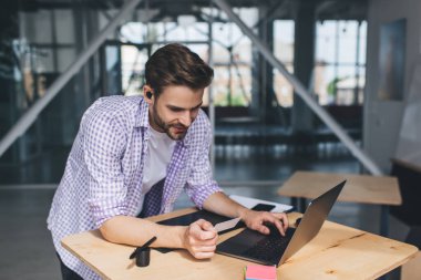 Milenyum Kafkas işadamı kartvizitten bir şey arıyor ve dizüstü bilgisayar kullanıyor. Modern başarılı adam kavramı. Açık uzay ofisindeki masada kablosuz kulaklıklı genç adam.