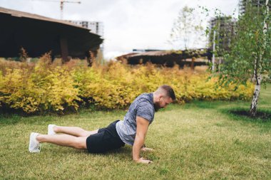 Spor kıyafetleri içinde genç sakallı bir adamın, binaların yakınındaki yeşil çimenli çayırlarda yoga yaparken Bhujangasana 'da yaptığı tüm vücut manzarası.