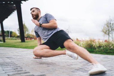 Aktif giyimli kararlı sakallı erkek sporcu, bulutlu bir günde parkta kaldırım kenarında spor yaparken yan tahta egzersizi yapıyor.