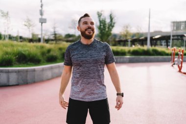Aktif giyimli genç, sakallı, sporcu kılıklı, güneşli yaz günlerinde spor sahasında durup başka tarafa bakan pozitif bir sporcu.