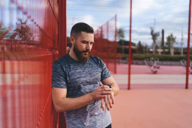 Spor kıyafetli sakallı erkek sporcu kırmızı ağ çitin yanında duruyor ve spor eğitmenini beklerken kol saatine bakıyor.