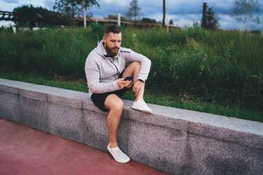 Spor giyimli, taş sınırında oturan ve parkta dinlenirken cep telefonunu karıştıran sakallı bir erkek sporcunun tüm vücudu.