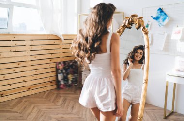 Arka planda uzun kıvırcık saçlı, uzun kıvırcık saçlı, gülümseyen bir kadın sabah uyandıktan sonra aynanın karşısında dikiliyor.