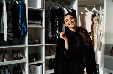 Tarz sahibi kadın gülümseyip kameraya bakarken modern kıyafetleri seçiyor ve evde çeşitli kıyafetler ve topuklularla dolaşıyor.