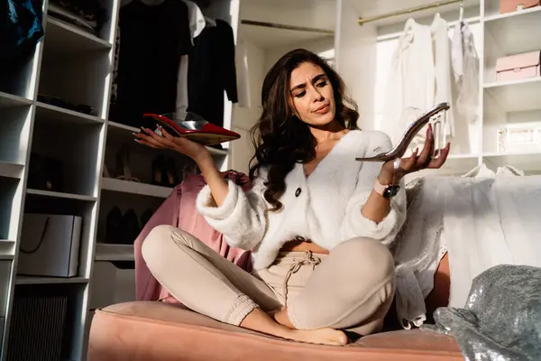 stock image Uncertain young woman in casual clothes sitting on comfortable couch trying to decide between shoes against closet in modern flat