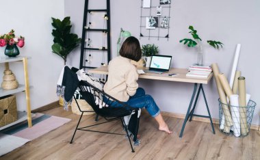 İskemlede oturan ve evdeki oturma odasındaki ağ kitabını kullanarak uzak bir projede çalışan sıradan giyinen isimsiz kadın serbest yazar.