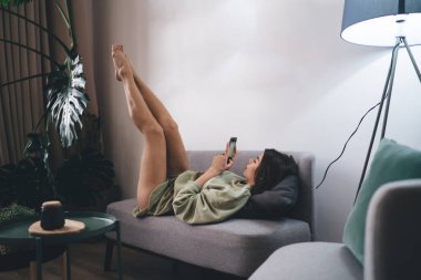 Kanepede uzanan ve evde dinlenirken akıllı telefon kullanan tüm vücudu gören genç bir kadın.