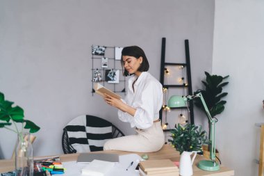 Beyaz bluz içinde kitap okurken gülümseyen ve yaratıcı çalışma alanında masa başında oturan genç ve konsantre bir esmerin yan görüntüsü.