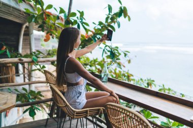 Hasır yüksek taburede oturan ve Tayland 'a yaz gezisi sırasında tropikal tatil yerindeki kafede cep telefonuyla kendi portresini çeken kimliği belirsiz genç bir kadının yan görüntüsü.