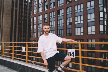 Modern binanın çatısında dikilen ve kameraya bakarken korkuluklara tutunan şık saç kesimi olan ciddi ve genç sakallı bir adam.