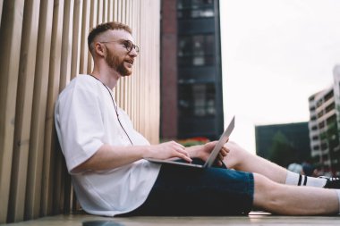 Sıradan kıyafetler ve gözlüklerle gülümseyen sakallı bir erkek öğrencinin ahşap bankta oturup dizüstü bilgisayarla uzaktaki bir caddeye bakarken görüntüsü.