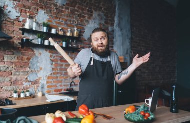 Kafası karışmış şef önlüklü bir adamın portresi. Elinde oklavayla kameraya bakıyor, omuzlarını silkiyor ve mutfakta taze sebzelerle tezgahın yanında duruyor.