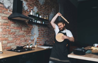 Beyaz tişörtlü, önlüklü, neşeli, tombul bir adam. Tahta kesme tahtasıyla savunuyor ve evdeki mutfakta tahta bir oklavayla tencereye vuruyor.