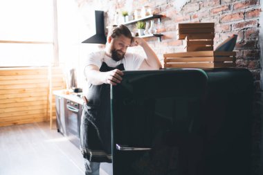 Beyaz tişörtlü ve önlüklü üzgün sakallı aşçı boş buzdolabına bakıyor ve evinde tuğla duvarla modern mutfakta iç çekiyor.