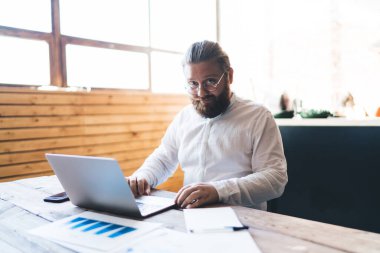 Gözlüklü, gülümseyen sakallı adam, elinde belgelerle oturmuş, netbook 'un klavyesine yazı yazarken modern kafede çalışıyor ve kameraya bakıyordu.