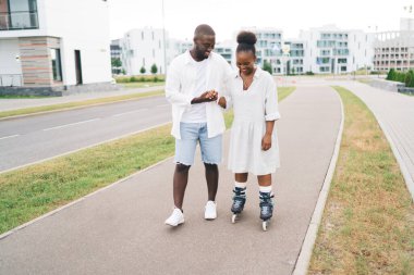 Hafta sonları asfalt kaldırımda yürürken kız arkadaşına paten kayarak yardım eden, gülümseyen Afrikalı Amerikalı erkek arkadaş.