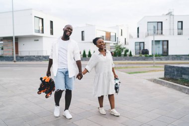 Genç Afrikalı Amerikalı erkek, hem paten taşırken hem de gün ışığında kaldırımda yürürken neşeli zenci kız arkadaşının elini tutuyor.