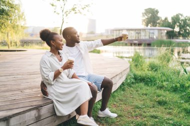Parkta, gölün kenarında tahta kaldırımda oturan Afro-Amerikalı bir kadın görülüyor. Elinde kahve fincanı tutan bir adam var.