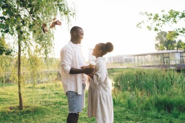 Yeşil ağaçların yanında el ele tutuşan ve birbirlerine gülümseyen Afro-Amerikan kadın ve erkeğin yan görüntüsü