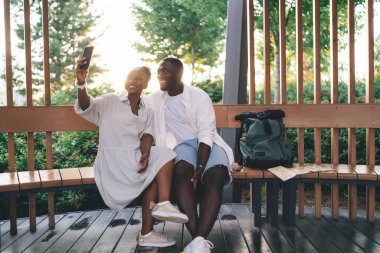 Sırt çantasıyla ahşap bankta oturmuş yeşil ağaçların ve çalıların fotojenik arka planına selfie çekerek gülümseyen Afrikalı Amerikalı sevgili ve kız arkadaş.