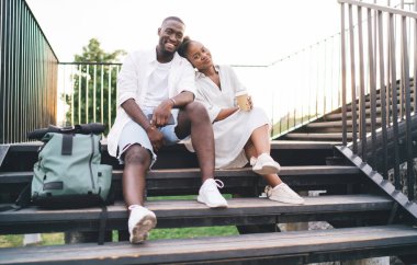 Yakışıklı siyah adam basamaklarda otururken Afro-Amerikalı çekici kız arkadaş kahve fincanı ile başını omzuna yaslıyor ve parkta kameraya gülümsüyor.