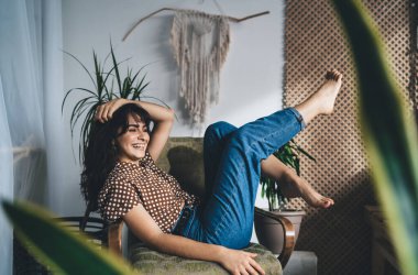 Neşeli, genç bir bayanın gündelik kıyafetleriyle koltuğa oturması, bacaklarını kaldırması ve elleriyle saçlarına dokunup başını çevirmesi.