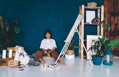 Çekici genç bayan sanatçı stüdyo zemininde oturmuş hobisinin keyfini çıkartıyor. Yaratıcı ve yetenekli esmer kadın sehpada resim yaratıyor. Petrolle çizim yapmayı öğrenmek