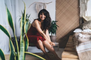 Yatak odasının yanındaki rahat koltukta oturan, bacağa dokunup kameraya bakan, çıplak ayaklı, çekici bir kadın.