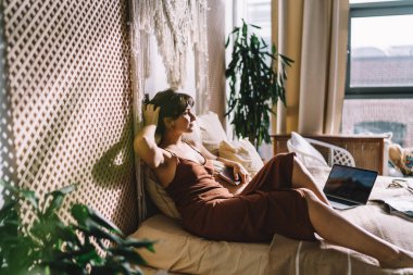 Düşünceli kadın serbest yazar netbook 'un yanında dinlenirken rahat bir yatakta otururken ve başka yere bakarken.