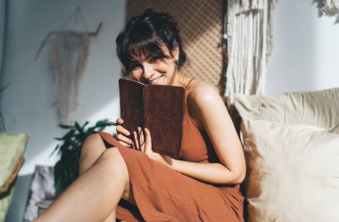 Güneşli bir günde elinde kalem ve defterle yatak odasında oturmuş kameraya gülümseyen etnik bir kadın.