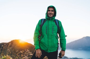 Yeşil yağmurluklu mutlu genç erkek yürüyüşçü çimenli dağda duruyor ve tatilde dağlık araziye giderken kameraya bakıyor.