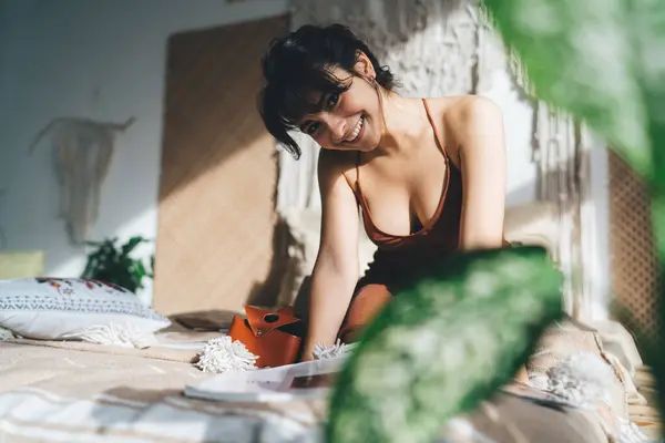 stock image Cheerful young woman in casual clothes looking at camera and smiling while siting on bed and reading magazine in bedroom at home on weekend day