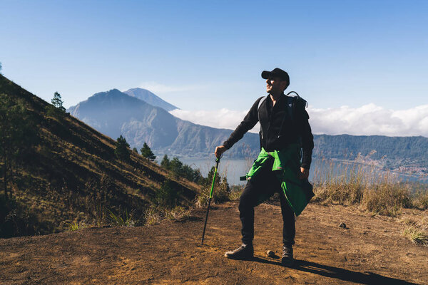 Полный набор содержания молодой мужчина турист в повседневной одежде и кепке опираясь на пешеходный полюс и глядя в сторону, стоя на смотровой площадке над озером во время поездки возле горы Батур