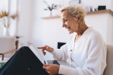 Koltukta oturan, bilgisayardaki verilere gülümseyen ve uzaktan kumanda sırasında parmaklarıyla işaret eden orta yaşlı kadın serbest çalışanın yan görüntüsü