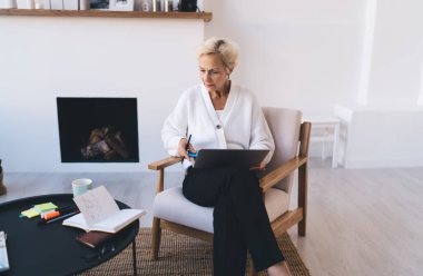 Masada oturmuş dizüstü bilgisayardaki bilgileri okurken not defterindeki notlara bakan orta yaşlı kadın.