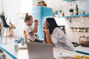 Beyaz tişörtlü mutlu genç annenin modern mutfakta elinde mandalinalarla oturmuş birbirine bakan kızına gülümsemesi.