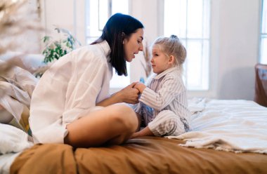 Yatakta rahat bir battaniyeyle oturan genç bir kadının yan görüntüsü. Uyku kıyafetleri içindeki bir çocukla konuşuyor ve birbirlerine bakıyor.